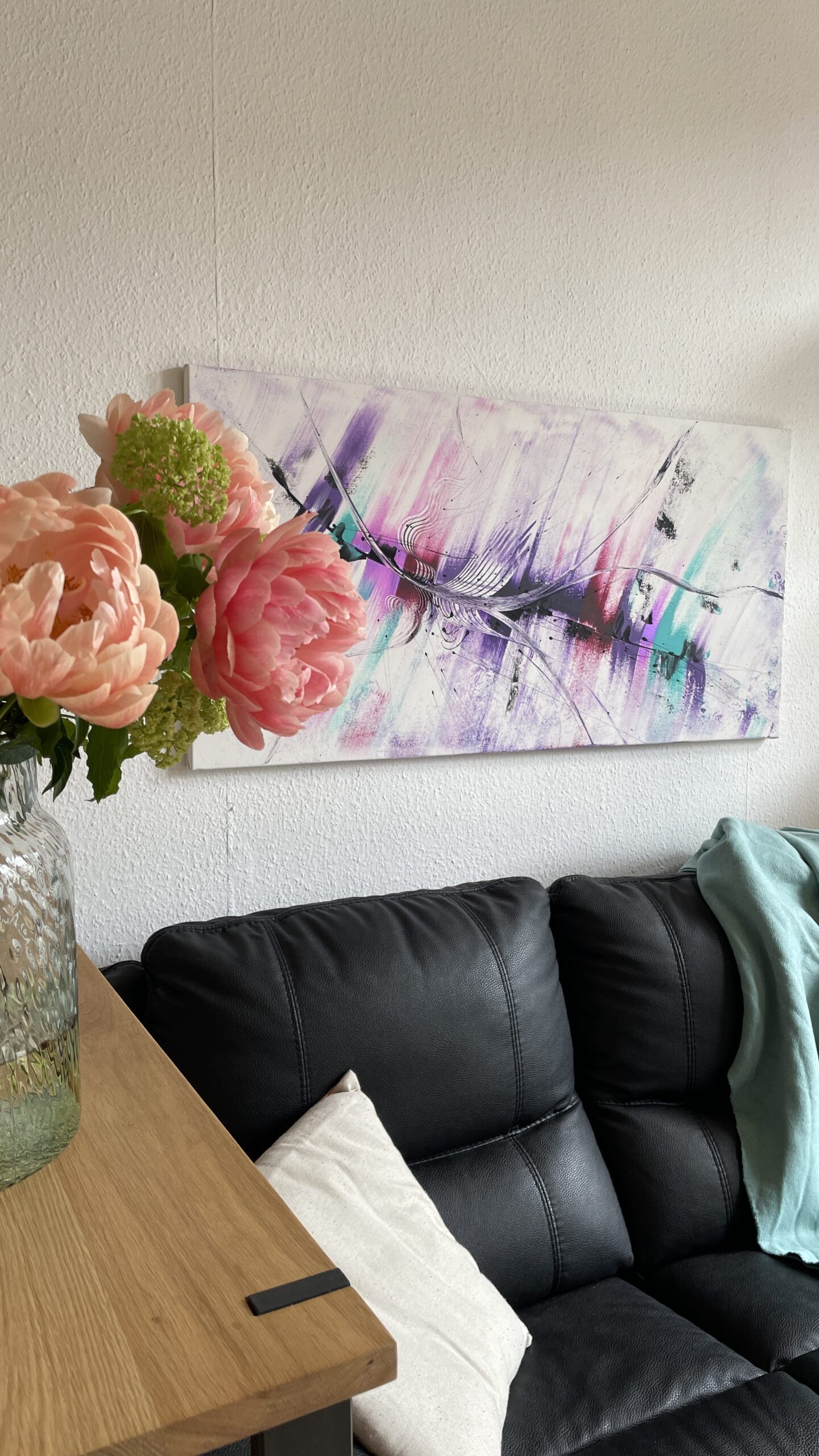 Abstraktes Acrylgemälde auf Leinwand. Es ist 120 cm x 60 cm groß in Lila, weiß und türkis. Das schöne Acrylgemälde hängt an einer Wohnzimmerwand über einer schwarzen Ledercouch. Es sind auch Pfingstrosen zu sehen.