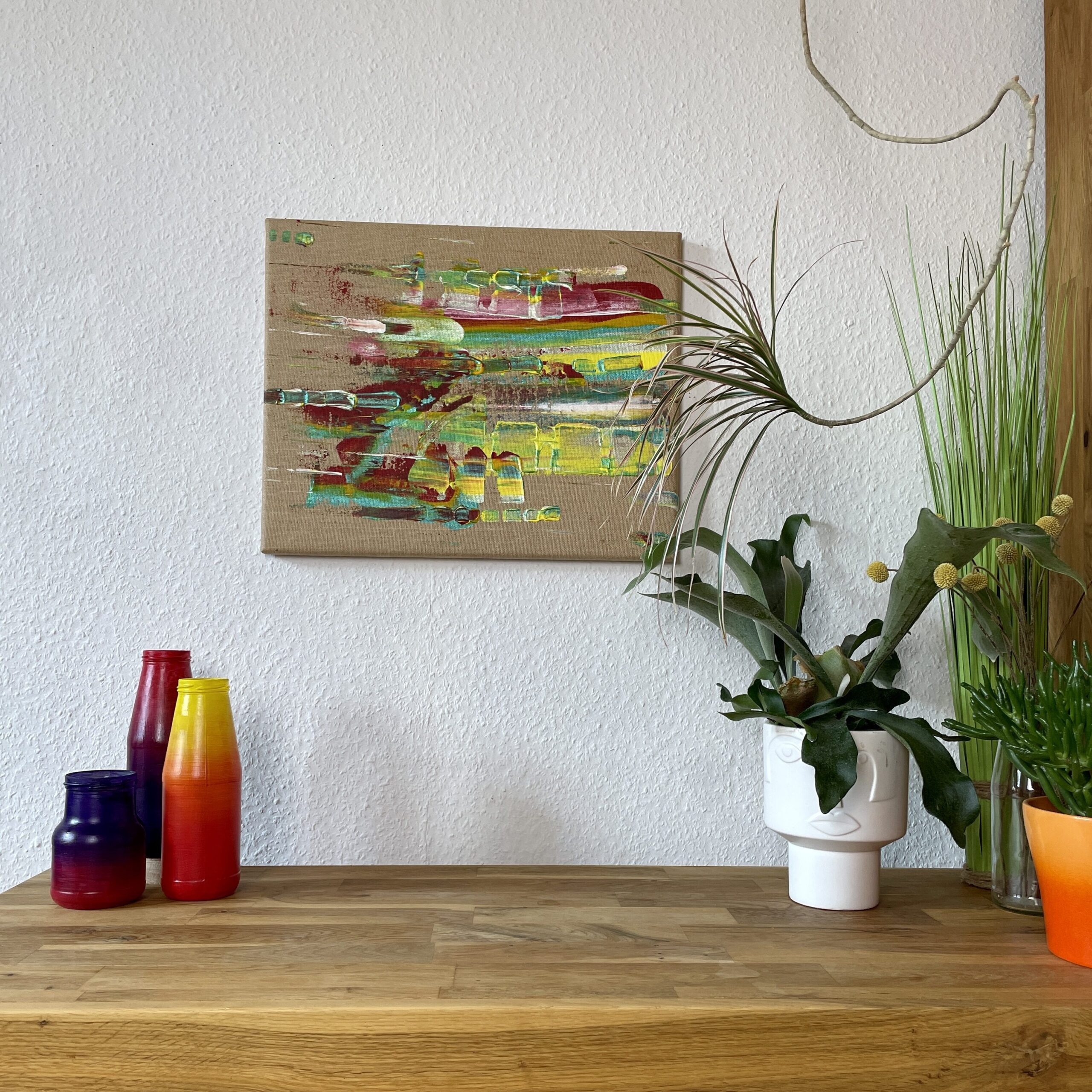 Tipps Kauf von Kunstwerken: Abstraktes buntes Acrylgemälde auf naturfarbener Leinwand handgemalt mit der Spachteltechnik. Es hängt an einer Wohnzimmerwand über einem Sideboard, auf dem bunte Gefäße und ein Geweihfarn in einem Keramiktopf stehen. Das Sideboard ist aus geöltem Eichenholz.