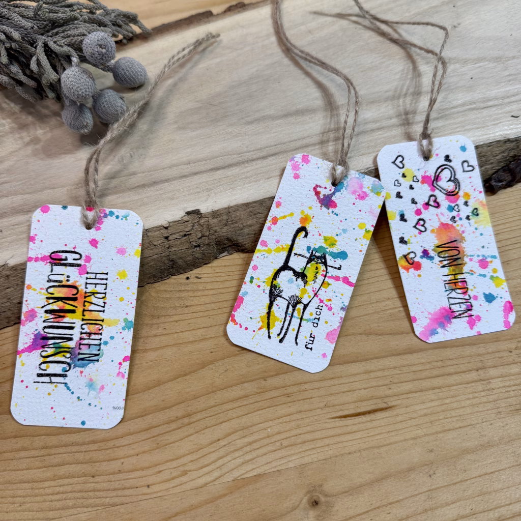 Handgefertigte bunte GeschenkanhĂ€nger mit SprĂŒchen wie âHerzlichen GlĂŒckwunschâ und âVon Herzenâ sowie einer Katze im Aquarell-Spritzstil, dekorativ arrangiert auf einem Holztisch.