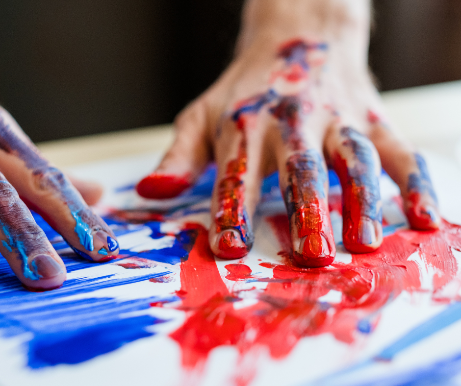 Erholen und zur Ruhe kommen mit malen. Zum Beispiel mit Fingerfarbe auf Papier. Malen kann zum Stressabbau helfen. Auf dem Foto siehst du zwei Hände, die mit hellroter und königsblauer Farbe auf Papier malen.